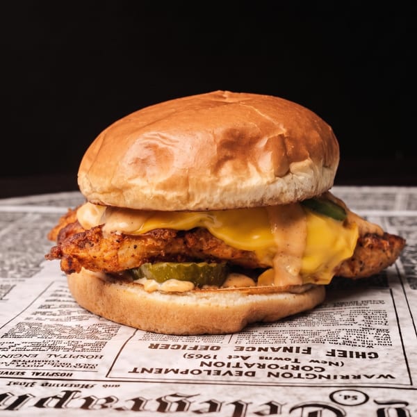 Crispy chicken sandwich with melted cheese, sauce, and pickles on a newspaper-lined surface.