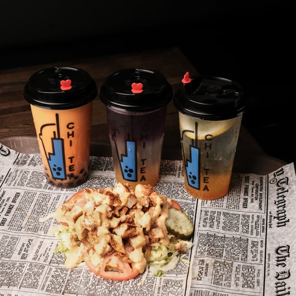 Three Chi Tea drinks and a chicken salad with tomatoes and pickles on newspaper-print paper.