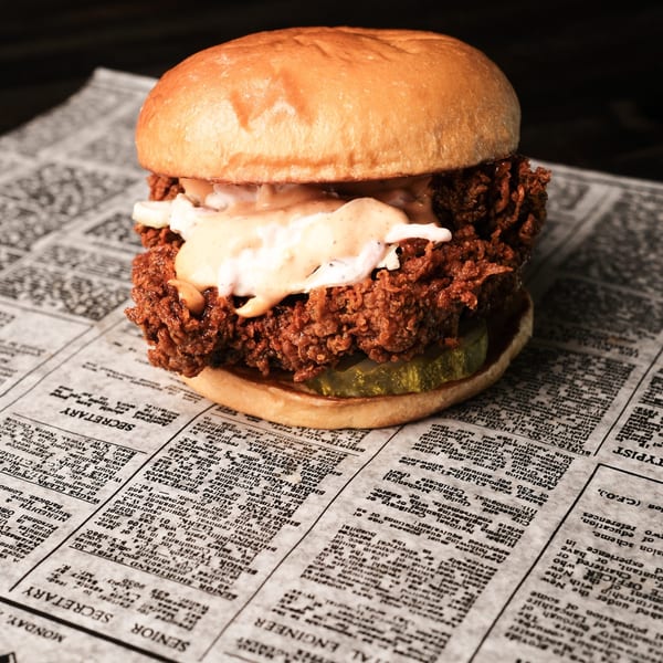 A crispy fried chicken sandwich with sauce and pickles rests on a newspaper.