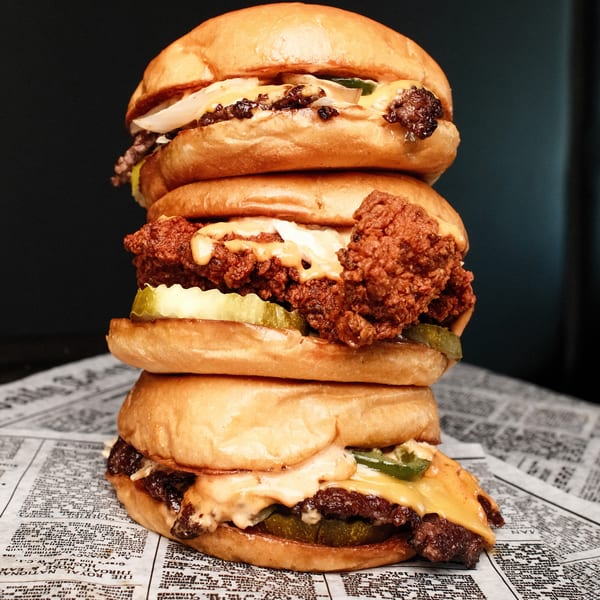 A towering stack of three burgers, including beef and fried chicken, on newspaper-patterned paper.