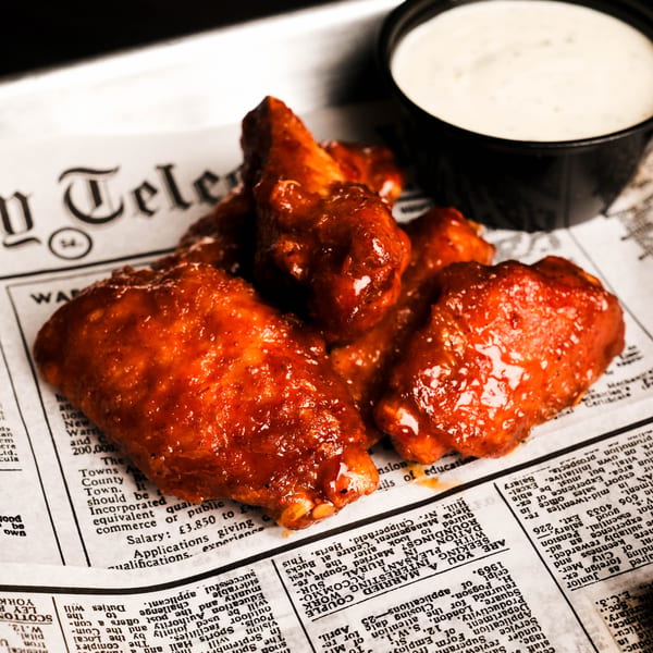 Glazed chicken wings and a side of ranch dressing served on newspaper-lined tray.