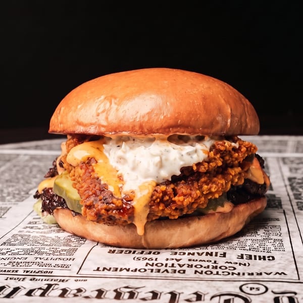 Large fried chicken burger with cheese, pickles, creamy white sauce, and orange sauce on a newspaper.