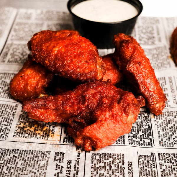 Spicy red chicken wings and a cup of ranch dressing on newspaper-lined tray.