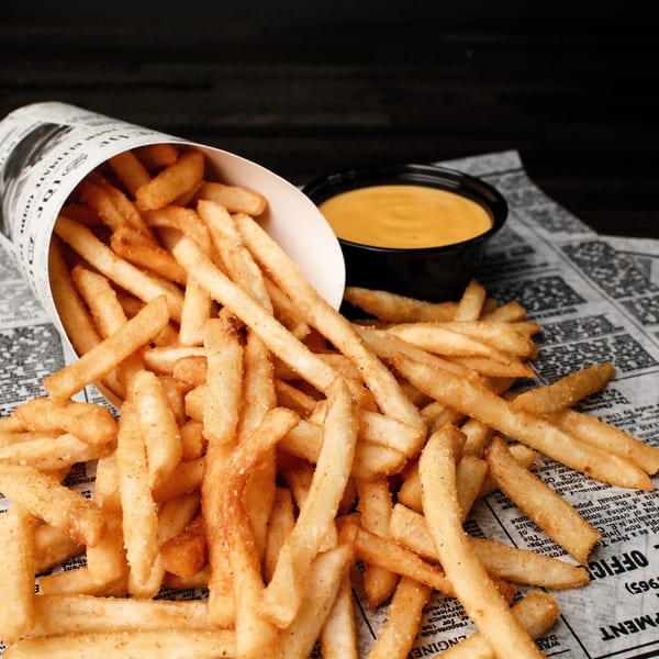 Crispy golden french fries spilling from a paper cone onto newspaper, with a side of cheese sauce.
