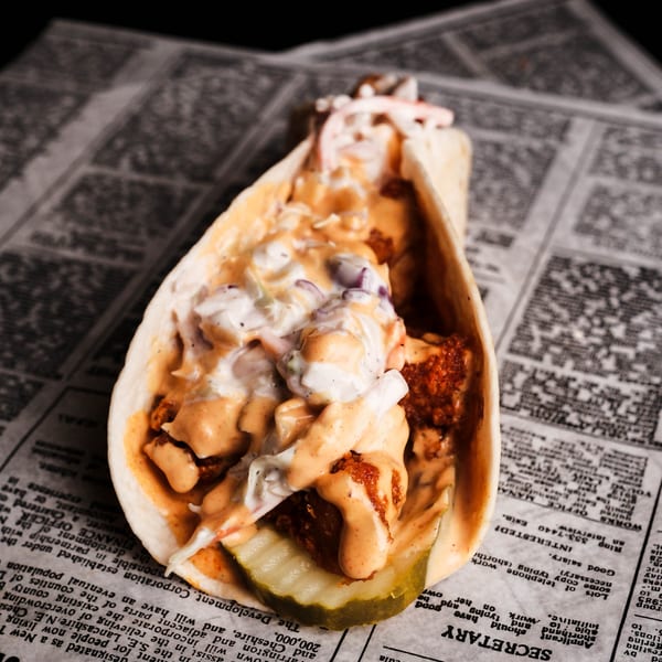 A fried chicken taco with coleslaw, sauce, and a pickle slice, on a newspaper.